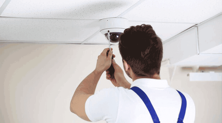 Um homem de camisa branca e suspensórios instalando uma câmera de segurança no teto
